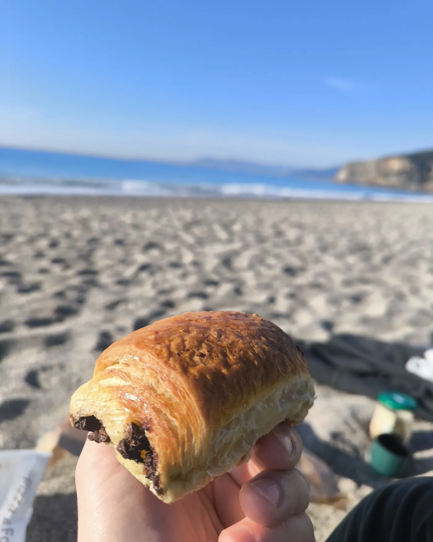 Una mano sostiene una medialuna de chocolate frente al mar, en la playa.