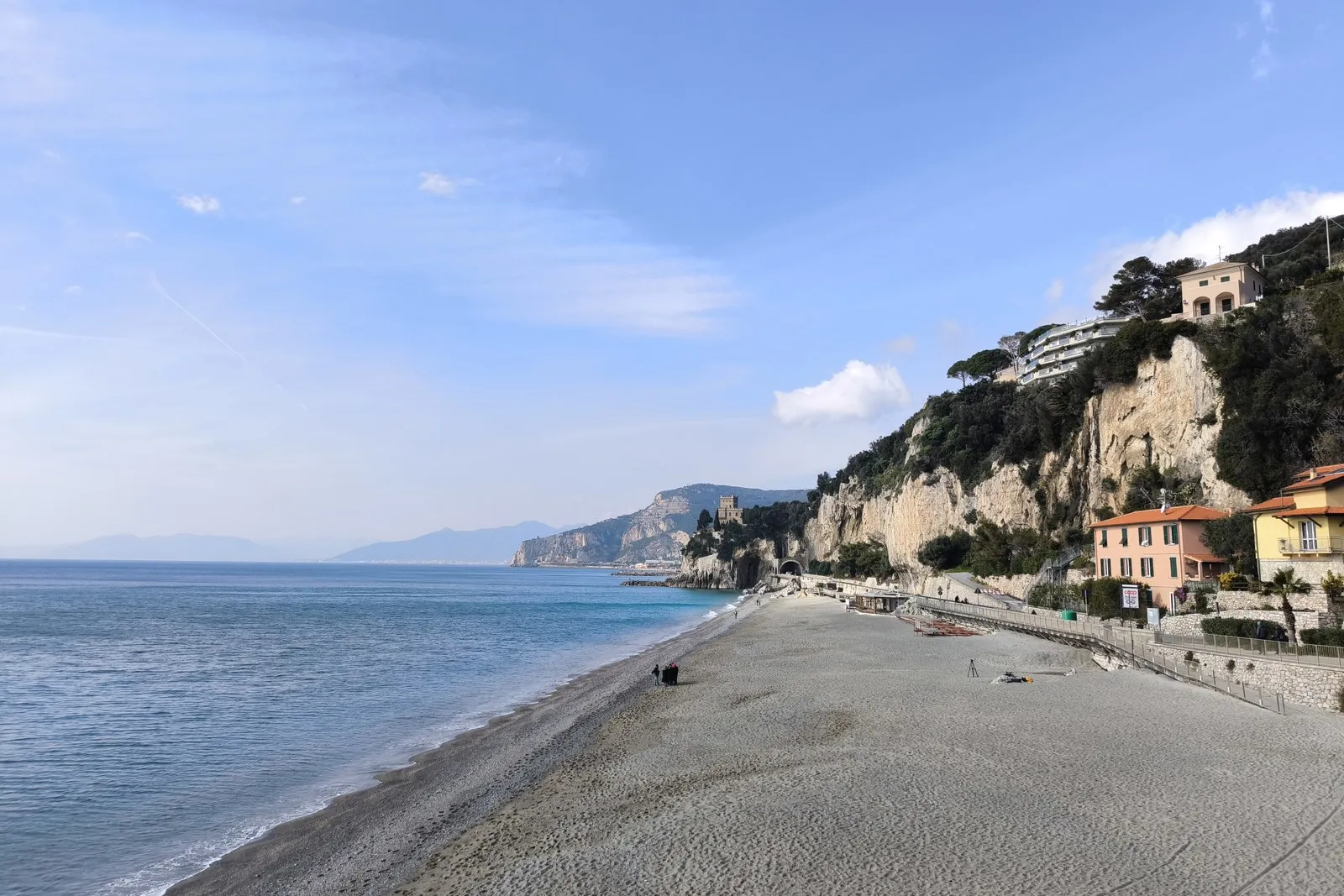 La playa de Finale Ligure con el mar calmo y un acantilado con casas a la derecha.